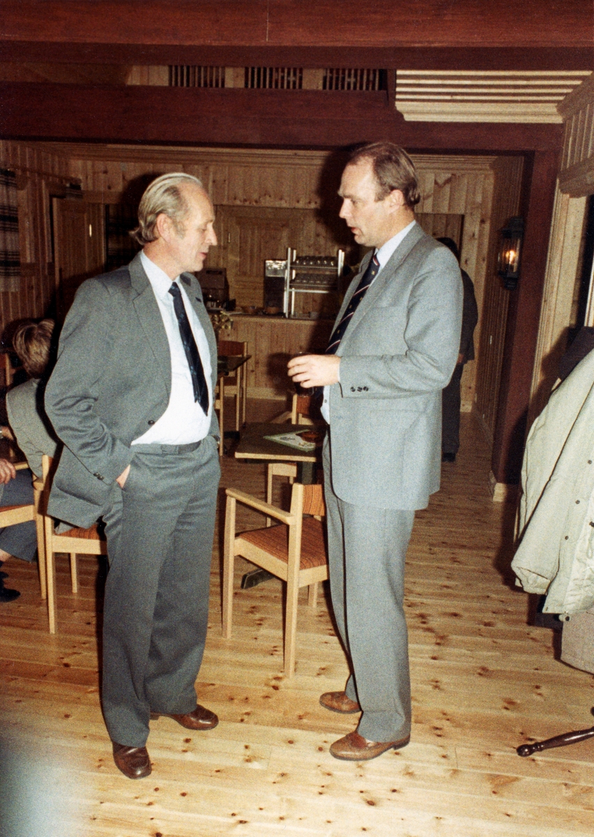 Kåre Brauten og Olav Støre i samtale i Våler Skurlags kontorbygning på Braskereidfoss i Solør.  Støre kom til Braskereidfoss i 1970, for å være med på å bygge Saga skogindustriers sponplatefabrikk.  I 1974 ble han knyttet til Skurlaget, sagbruksbedriften på nabotomta.   Støre var først teknisk sjef ved denne bedriften, men ble leder for sagbruket, først med tittelen "produksjonssjef" i 1977, seinere som "disponent".  Denne posisjonen hadde Støre de følgende ti årene.  I 1988 flyttet Støre tilbake til oppvekstmiljøet sitt i Trøndelag, der han overtok en familieeiendom og arbeidet som bankmann.  Kåre Brauten hadde en tilsvarende posisjon som den Støre hadde ved Våler Skurlag ved trelastbedriften van Severen i Namsos. Begge de nevnte sagbrukene inngikk i den tida da dette fotografiet ble tatt i Norske Skogs trelastdivisjon.