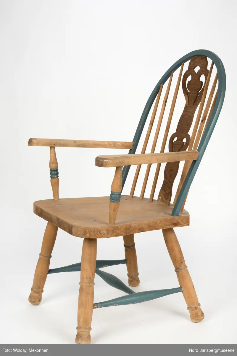"Budal"inspirert stol, med ryggstø og armelner. Ryggstø er formet av en blåmalt buet list som holdes oppe av seks vertikale trespiler. Den midter, og syvende, spilen er en bred, utskjært list med gjennombrutt mønster, stiliserte hjertemønster. Armlenene er buet utover, og ben og detaljer har dreid dekor.