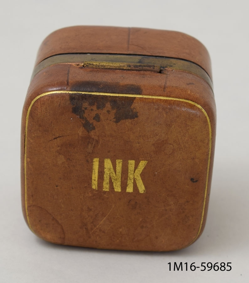 Läderbeklätt bläckhorn av metall beklätt med läder, i formen av en kub. Öppningsbart. Inuti ett bläckhorn av glas. Uanpå är det skrivet "INK".