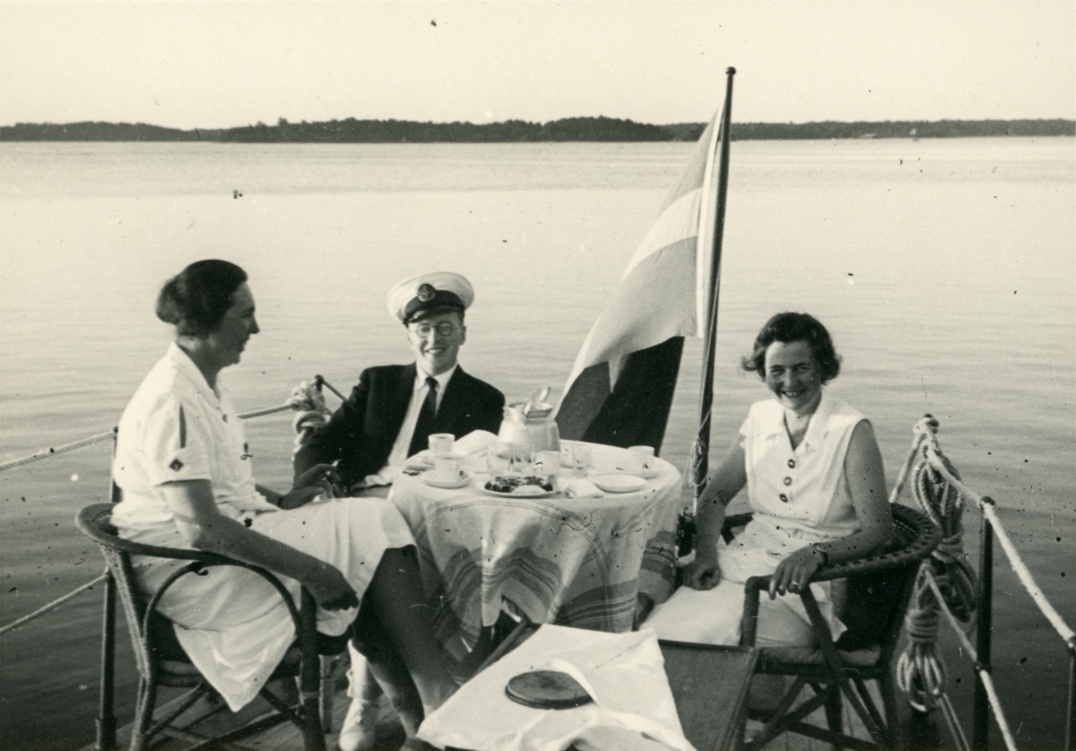 Två kvinnor och en man sitter vid ett dukat bord i aktern på en båt.
