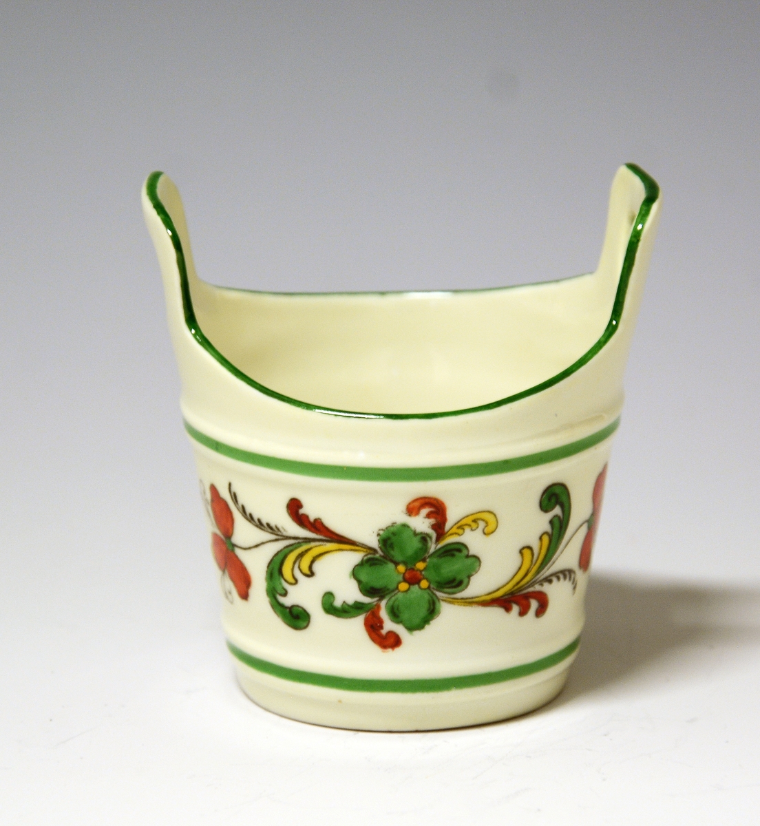 Liten kopp av porselen, formet som en smørstamp. Hvit glasur, også inni. På sidene rosemalt felt i sterke farger.
Modell: 1700
Dekor: 72679