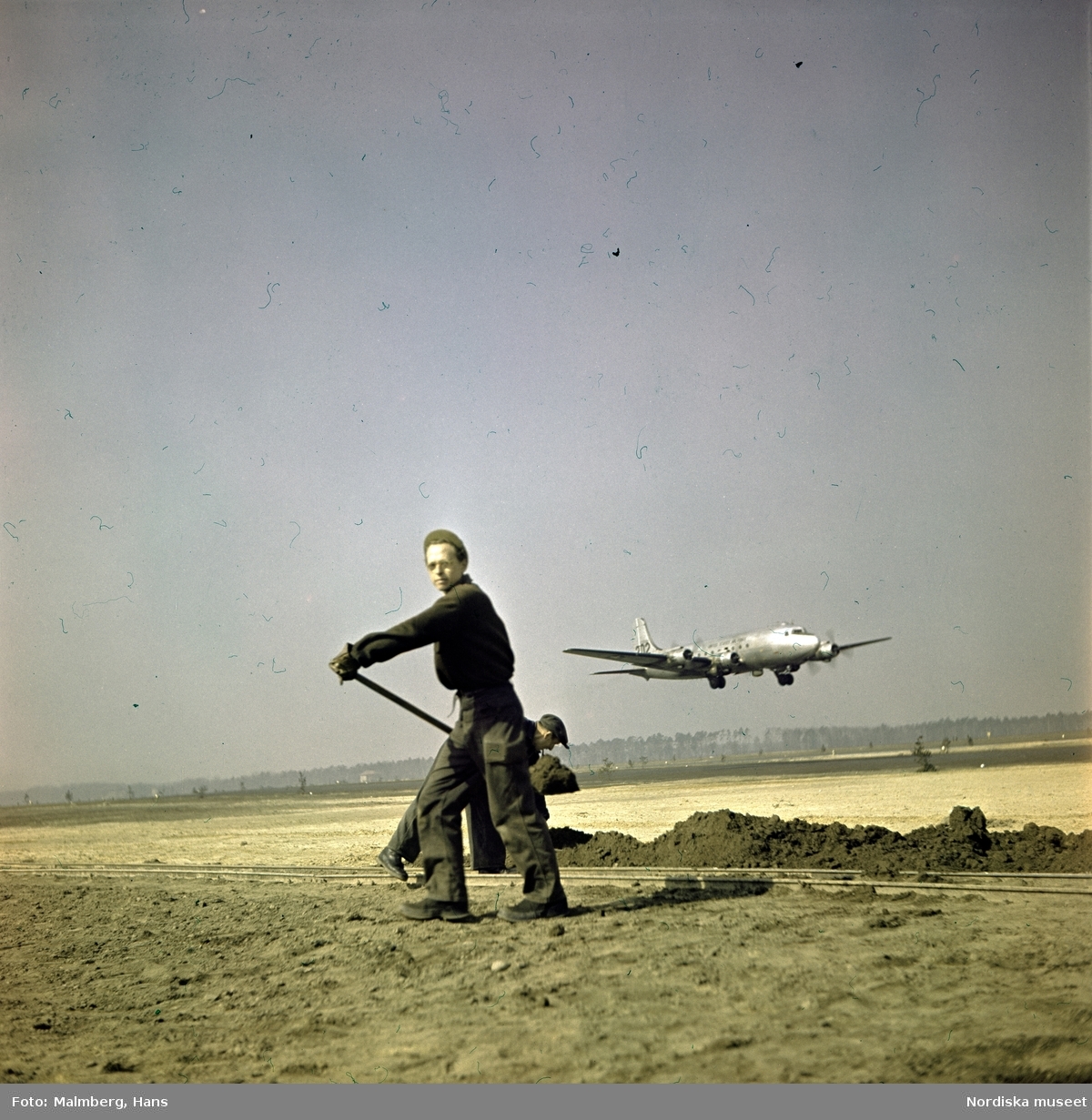 Berlinblockaden. En arbetare gräver på ett flygfält, i bakgrunden går ett transportflygplan in för landning.