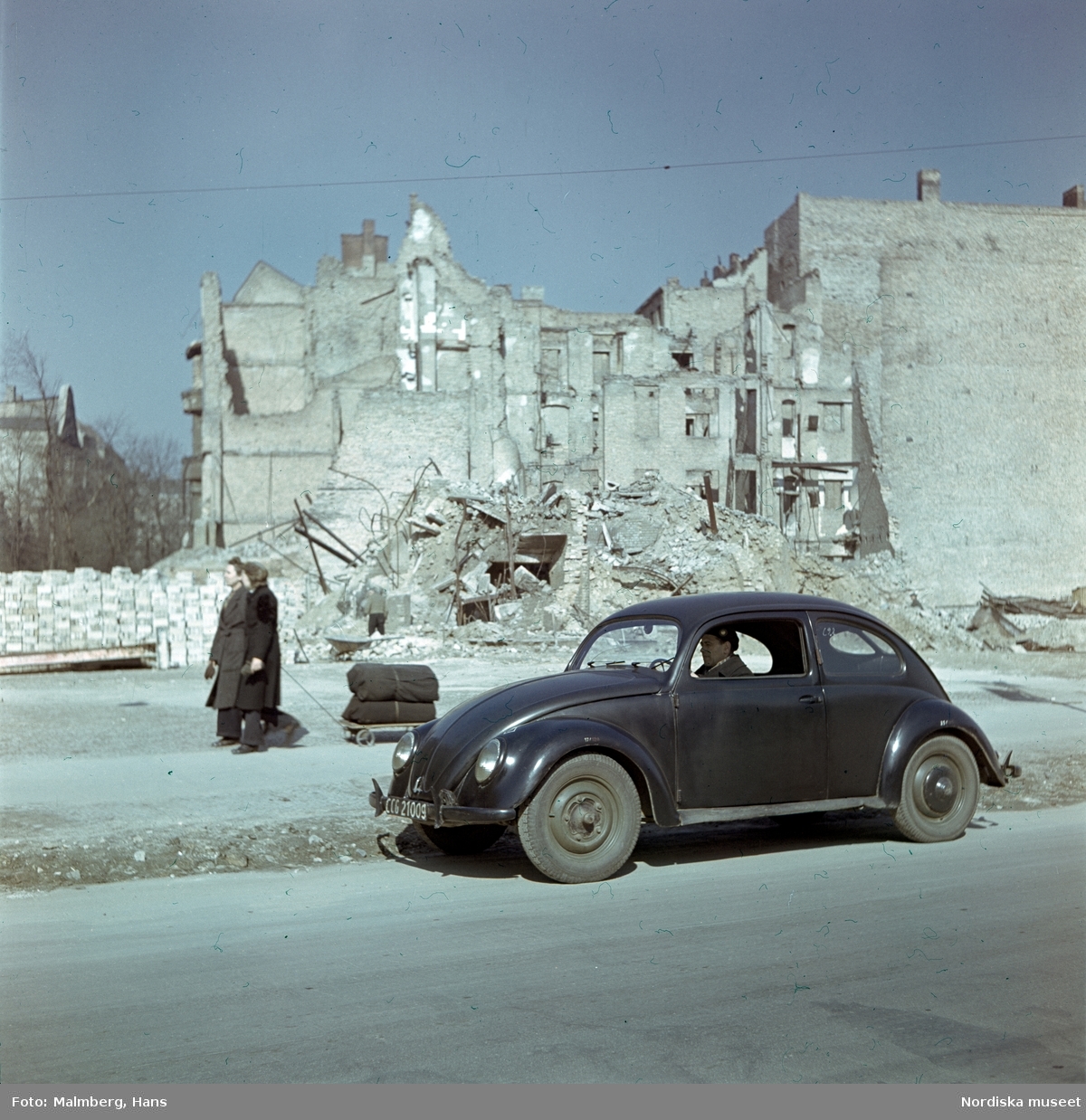 Berlin. En soldat ur de brittiska ockupationstrupperna kör en Volkswagen. I bakgrunden husruiner och två kvinnor med en kärra.