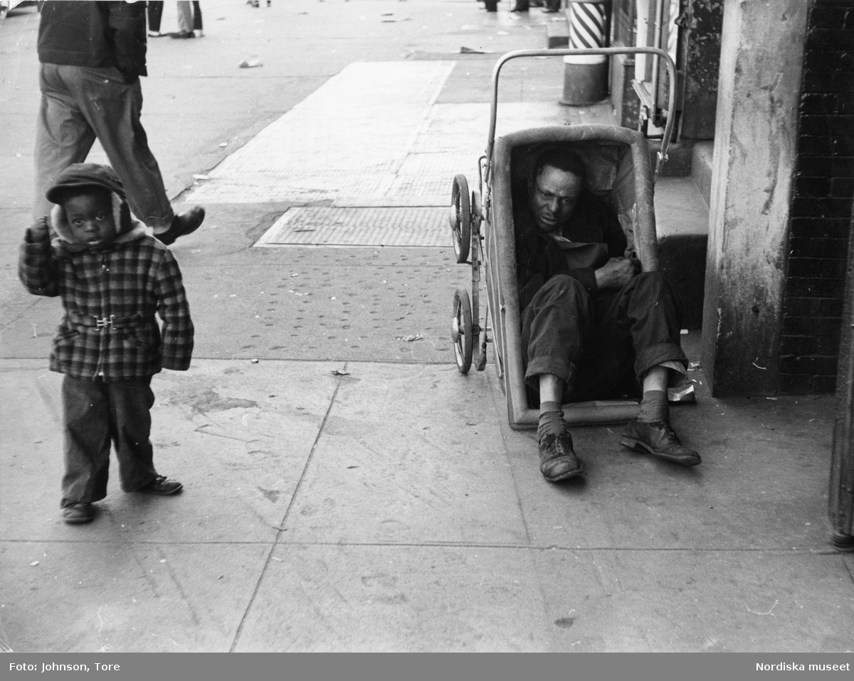 En pojke bredvid en barnvagn där en utslagen man sitter. Lenox Avenue, Harlem, New York, USA.