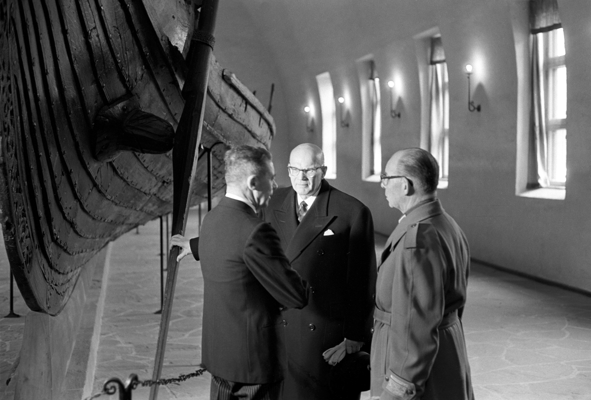 Finlands president Urho Kekkonen (i midten) er på statsbesøk i Norge. Her blir presidenten vist rundt av professor Bjørn Hougen (med ryggen til) på Vikingskipsmuseet på Bygdøy. Ukjent person til høyre.
