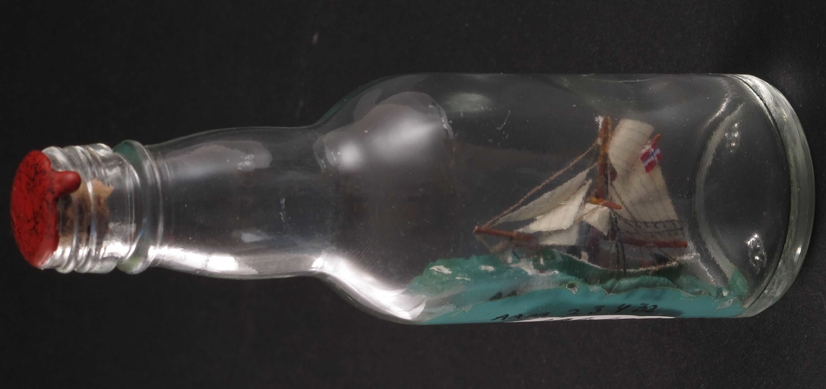 Miniatyr flaskeskute: Jakten "Gjøa".  Glass, klart. Kork. rød lakk. Innstøpt i bunnen:  MG  (Moss Glassverk)  12. Grønnmalt skrog, grønn sjø, norsk flagg.