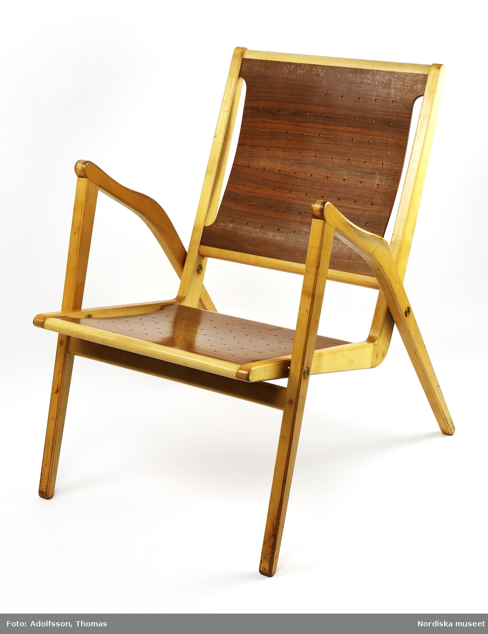 Stapelbar stol, karmstol, med stativ av klarlackad björk. Formpressad, perforerad sits och rygg av valnötsfaner. Sittdel och stativ sammansatta med mässingsbeslag av tryckknappstyp. Sammanfogning med slitsar som även ger en dekorativ verkan. Formgiven 1955 och sannolikt tillverkad samma år.
/Anna Arfvidsson Womack 2019-01-29