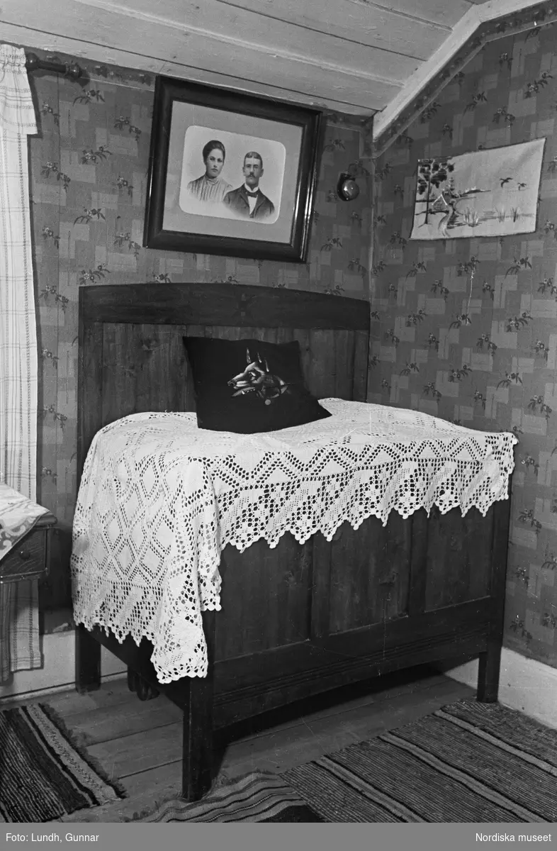 Interiör från ett statarhem. Säng med vitt överkast, kudde, porträtt på väggen ovanför sängen.