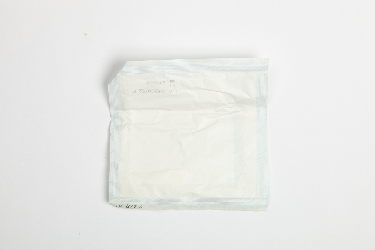 En steril kompress forseglet i en hvit papirpakning. Pakken er kvadratisk og flat. På oversiden står det hvilket innhold som er i papirpakningen.