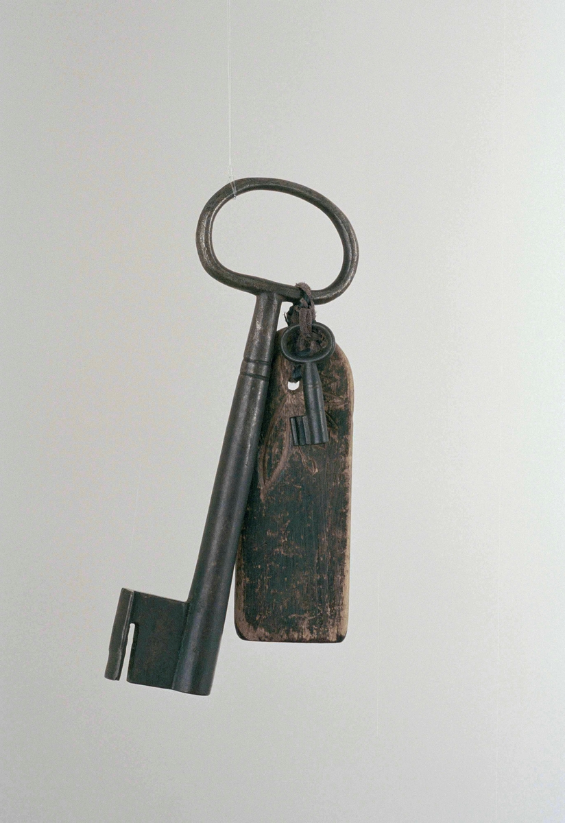 Snidad nyckelbräda(a) av furu med två tillhörande nycklar av järn, en stor(b), en liten(c). På ena sidan av brädan är ordet "nyckel" inristat, på andra sidan dekor.