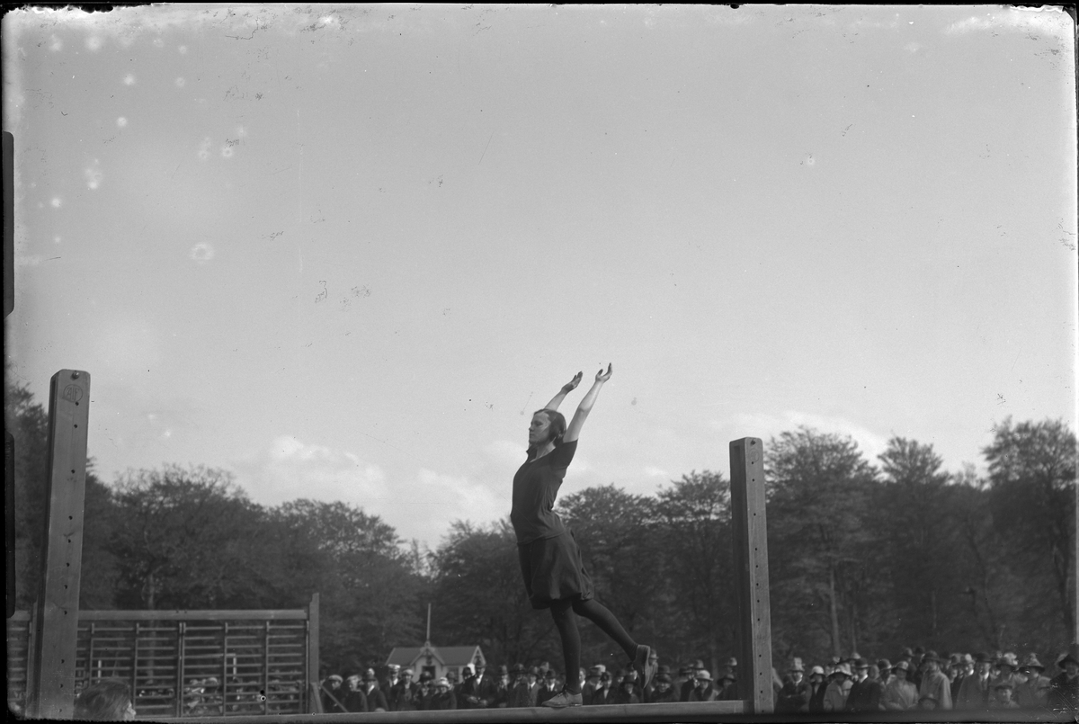 Kvinnlig gymnast går balansgång på en bom, runt om står åskådare. I fotografens egna anteckningar står: Danmarksresan, Balansgång.