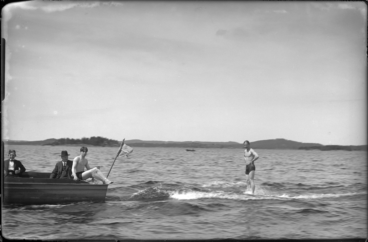 I en träbåt sitter en man och en pojke, båda kostymklädda. På båtens kant sitter en ung man i badkläder medan en annan man åker bakom båten. I fotografens anteckningar står det "Surfing, Axelsson".