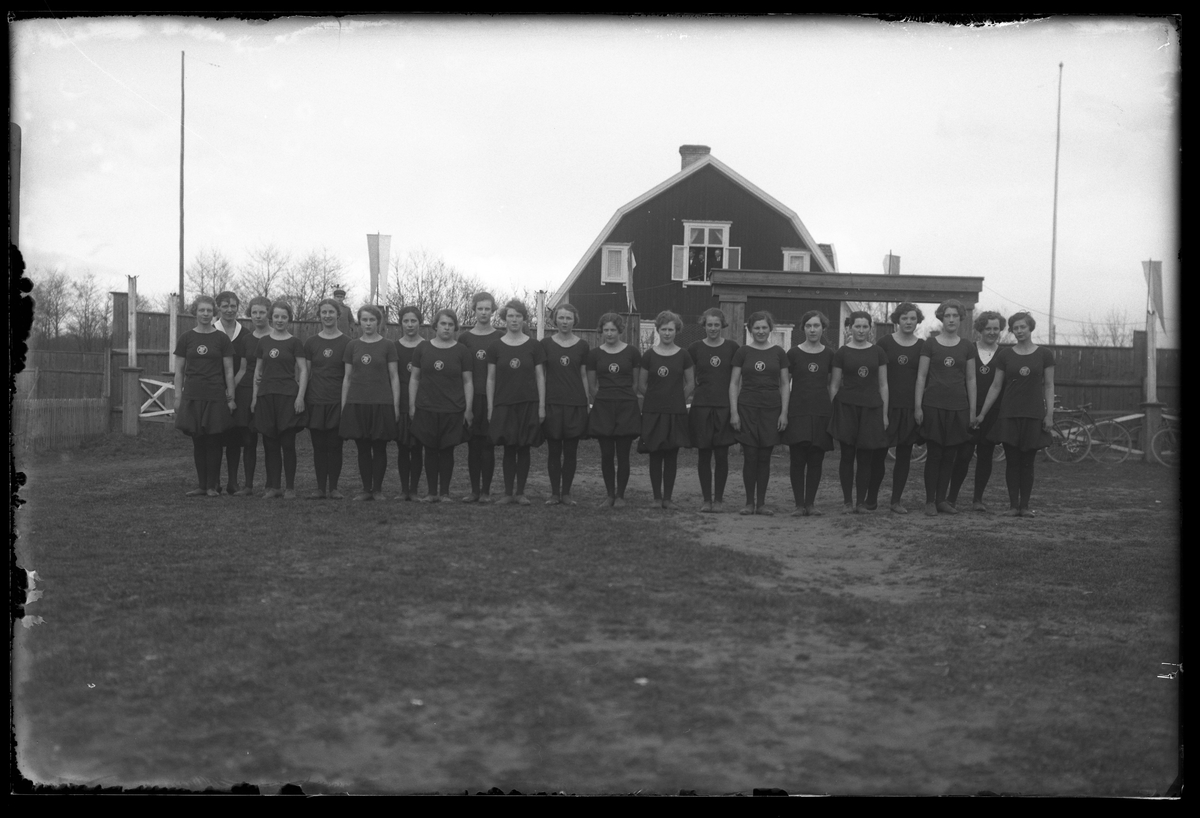 Gruppbild på Alingsås Idrottsförenings (AIFs) kvinnliga trupp i svarta dräkter. Bilden är tagen i samband med en gymnastikuppvisning. I övervåningens fönster på villan i bakgrunden tittar tre män ut.