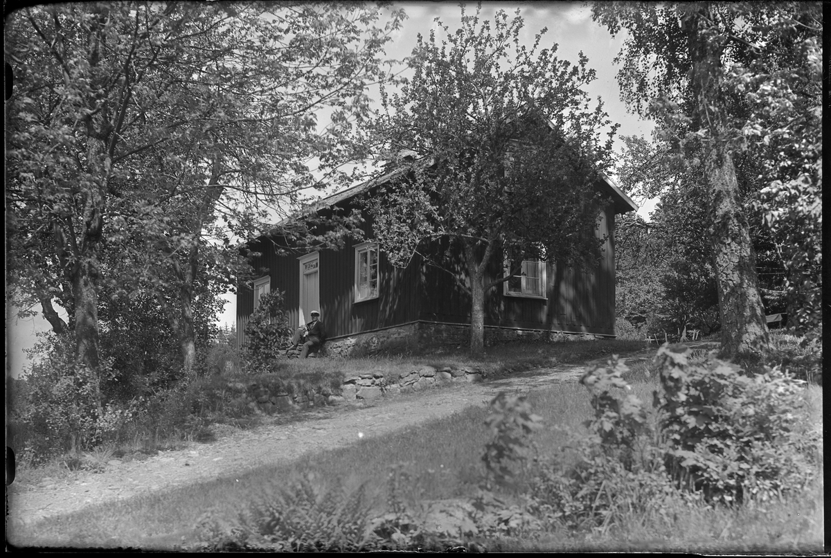 Ett mindre trähus uppe på en kulle med träd runt om. På trappen sitter en man med en hund, förmodligen Prisse, i knät. I fotografens egna anteckningar står det "Skoglunds hus".