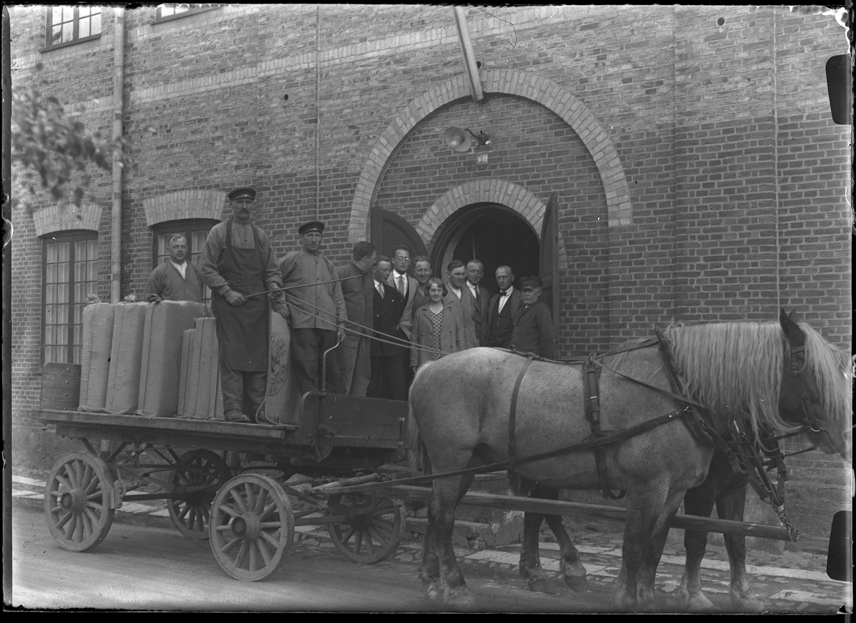 Två hästar med en vagn står utanför bomulls väveriet. Vagnen är lastad med textilier och på den står tre män. I dörröppningen till väveriet står packpersonalen. I fotografens anteckningar står det "drängarna och packarna".