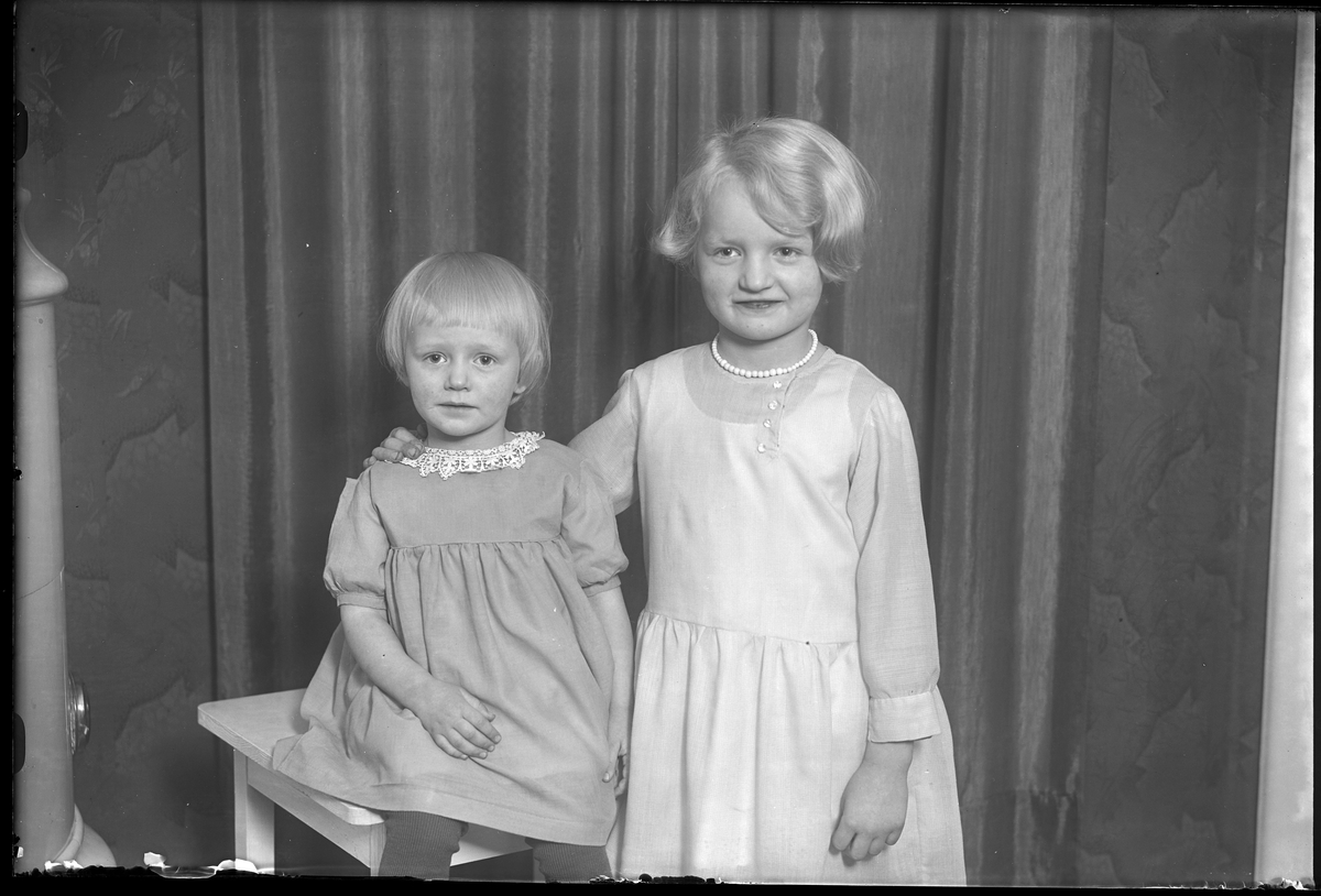 En flicka sitter på en pall medan hennes syster står bredvid och håller om hennes axlar. I fotografens egna anteckningar står det "Barnen Dahlén". Tolkat som Ann-Marie och Margit Dalén.