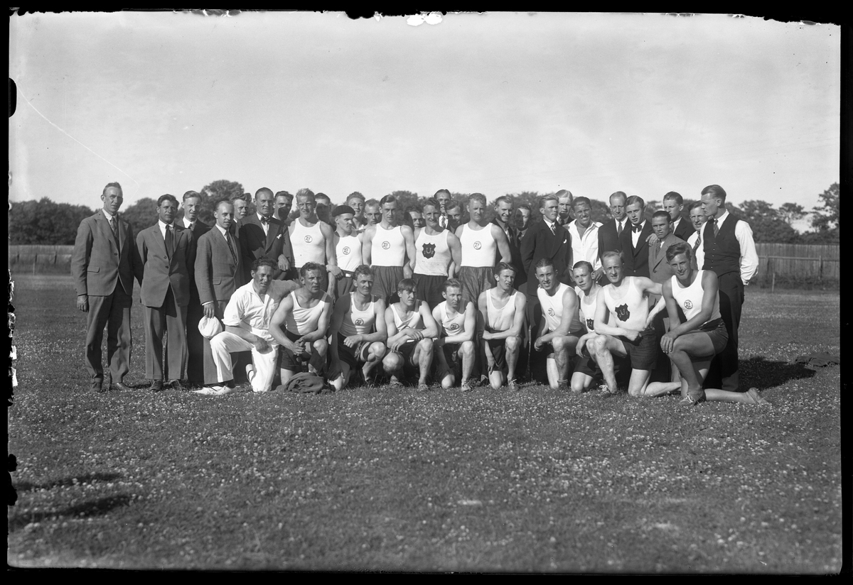 Gruppbild på uppradade män på en gräsplan. Vissa av männen bär idrottskläder medan andra bär kostym. I fotografens anteckningar står det "Ulricehamn - A.I.F."