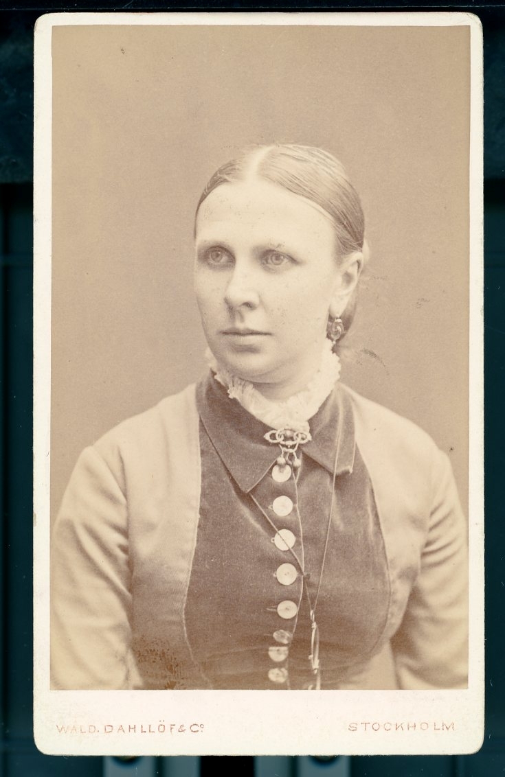 Kabinettsfotografi: bröstbild av Hanna Quennerstedt, brosch vid halsen och glasögon hängande i kedja runt halsen.