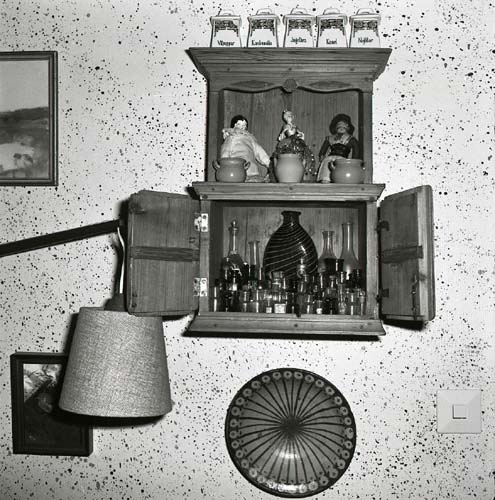 Ett väggskåp är fäst på en stänkmålad vägg. I skåpet står glasflaskor och dockor samt på skåpet står kryddburkar, 1984.