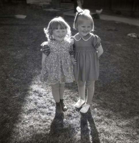 Två flickor iförda klänningar och knästrumpor poserar på en gräsmatta. Den ena flickan har en rosett i håret, 1950.