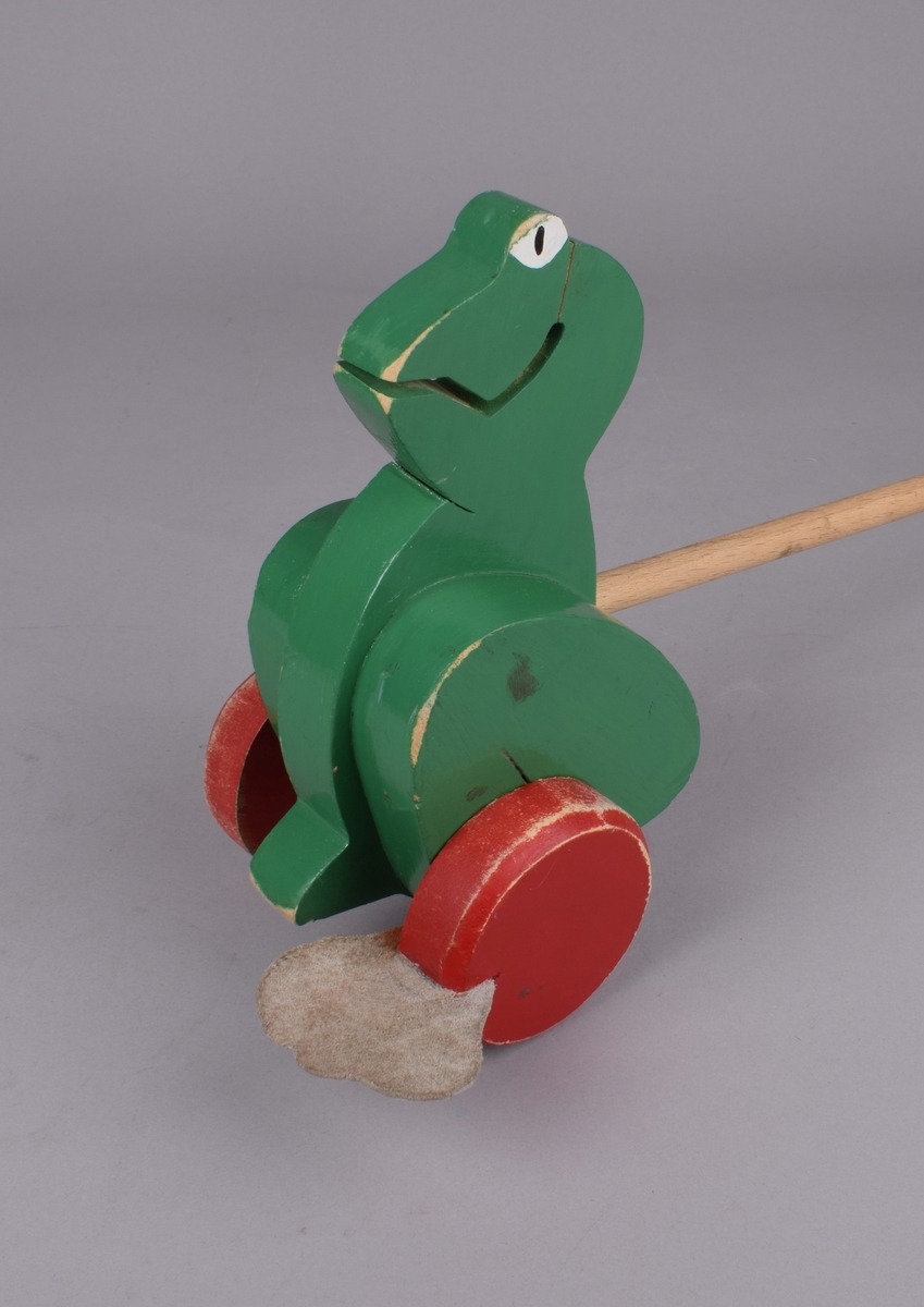 Utskåret froskefigur på to hjul med skinnføtter. Figuren er festet til en trepinne/håndtak med en kule i enden. Når figuren skyves går føttene på hjulene rundt slik at frosken "går".