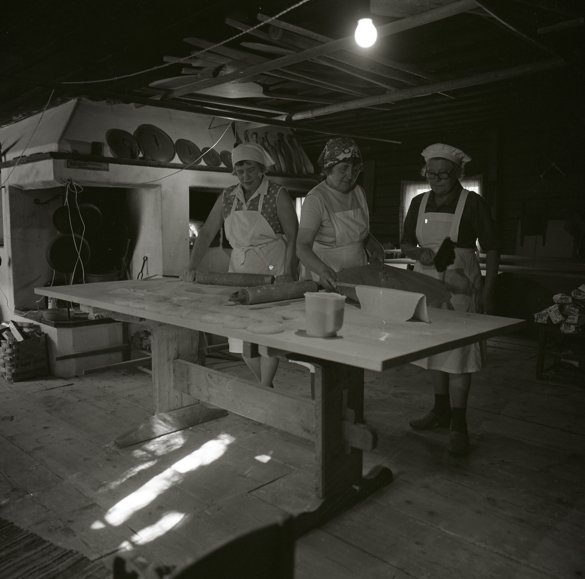 Tre kvinnor i förkläden och schaletter bakar tunnbröd vid ett bord. De använder kruskavlar, brödpinne och en brödspade. Ovanpå bakugnen bakom kvinnorna står grytlock och bakredskap samt vid öppna spisen hänger det grytor, Hembygdsbyn 7 juni 1979.