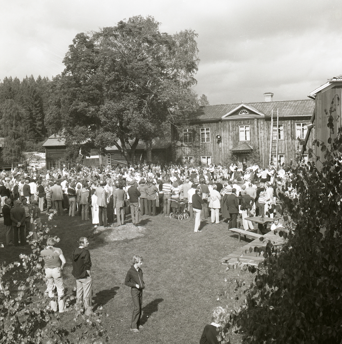 En grupp människor har samlats på en gård framför ett timrat hus under Rengsjöfesten 1980. Intill det timrade huset står det ytterligare några timrade byggnader.