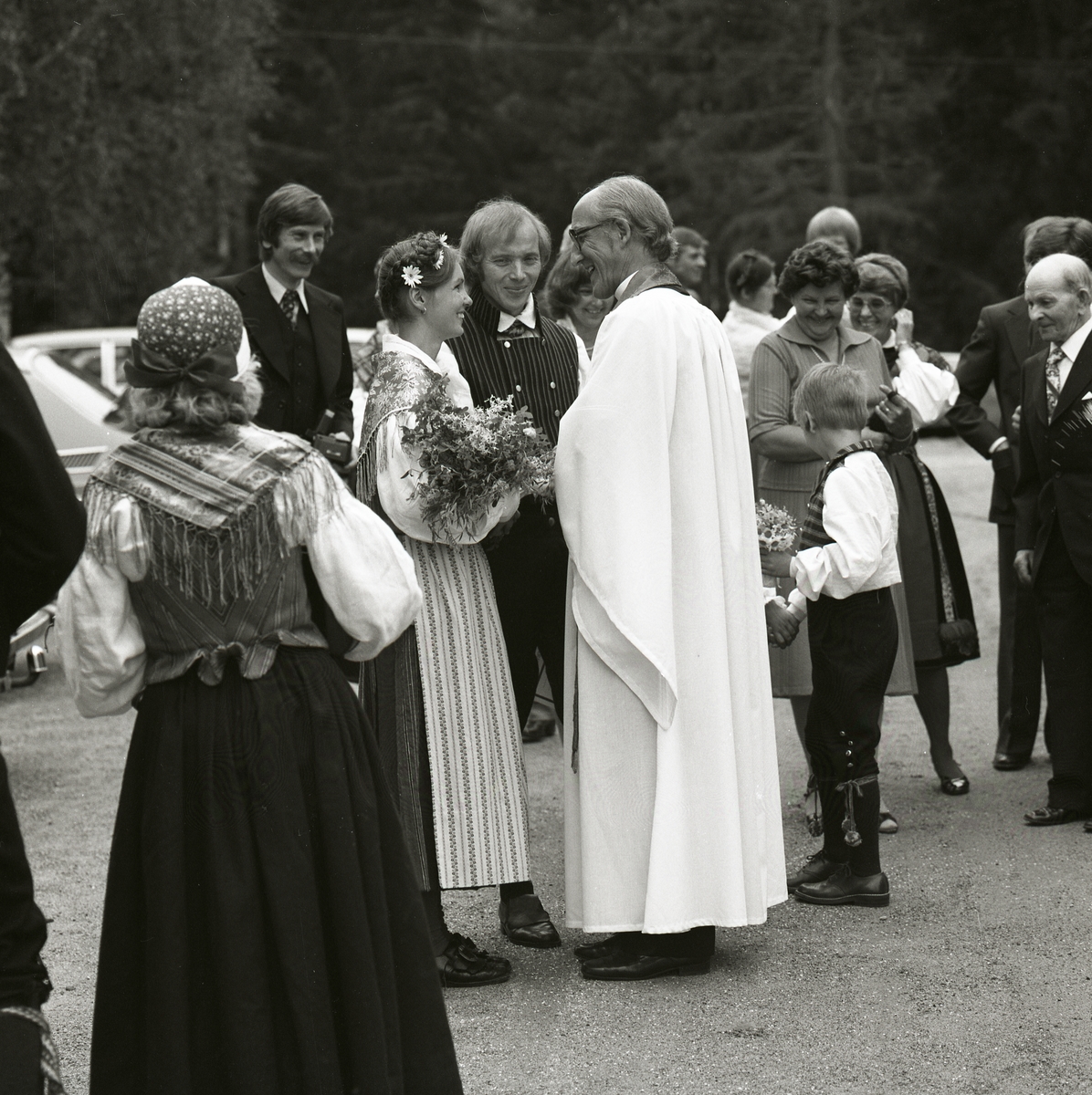 En grupp människor iklädda hembygdsdräkter och kostymer står runt ett brudpar. Bruden har blommor i sitt hår och talar med prästen som är klädd i vitt, 28 juni 1980.