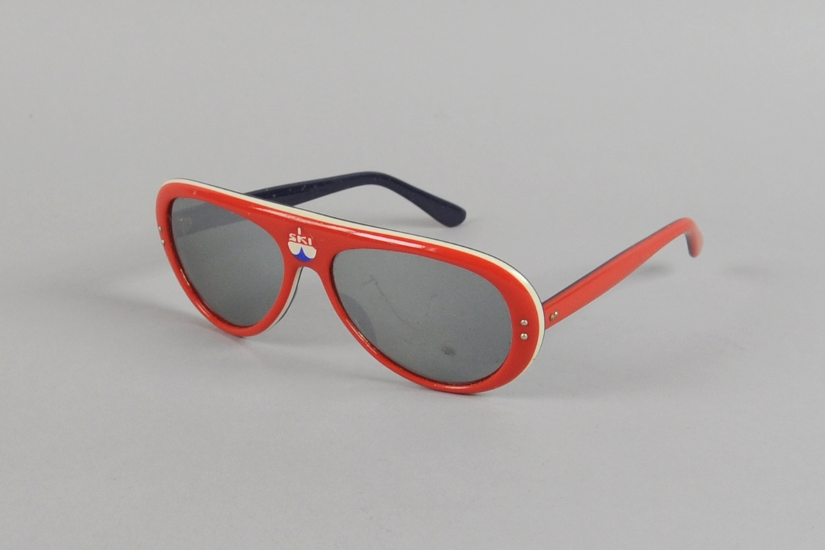 Solbriller med innfatning av plast i hvitt, rødt og blått og fargete glass.
Midt i brillen et merke med innskriften I SKI. Brillene ligger i et etui av skinnimitasjon merket Optisk Kompani A/S