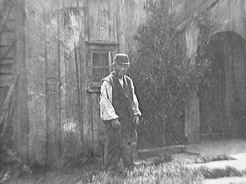 Ryggåsstuga med s k skunke vid entrén. Framför huset är marken belagd med stenhallar och där står en man i väst och keps. Bilden märkt "Carl Heintz".