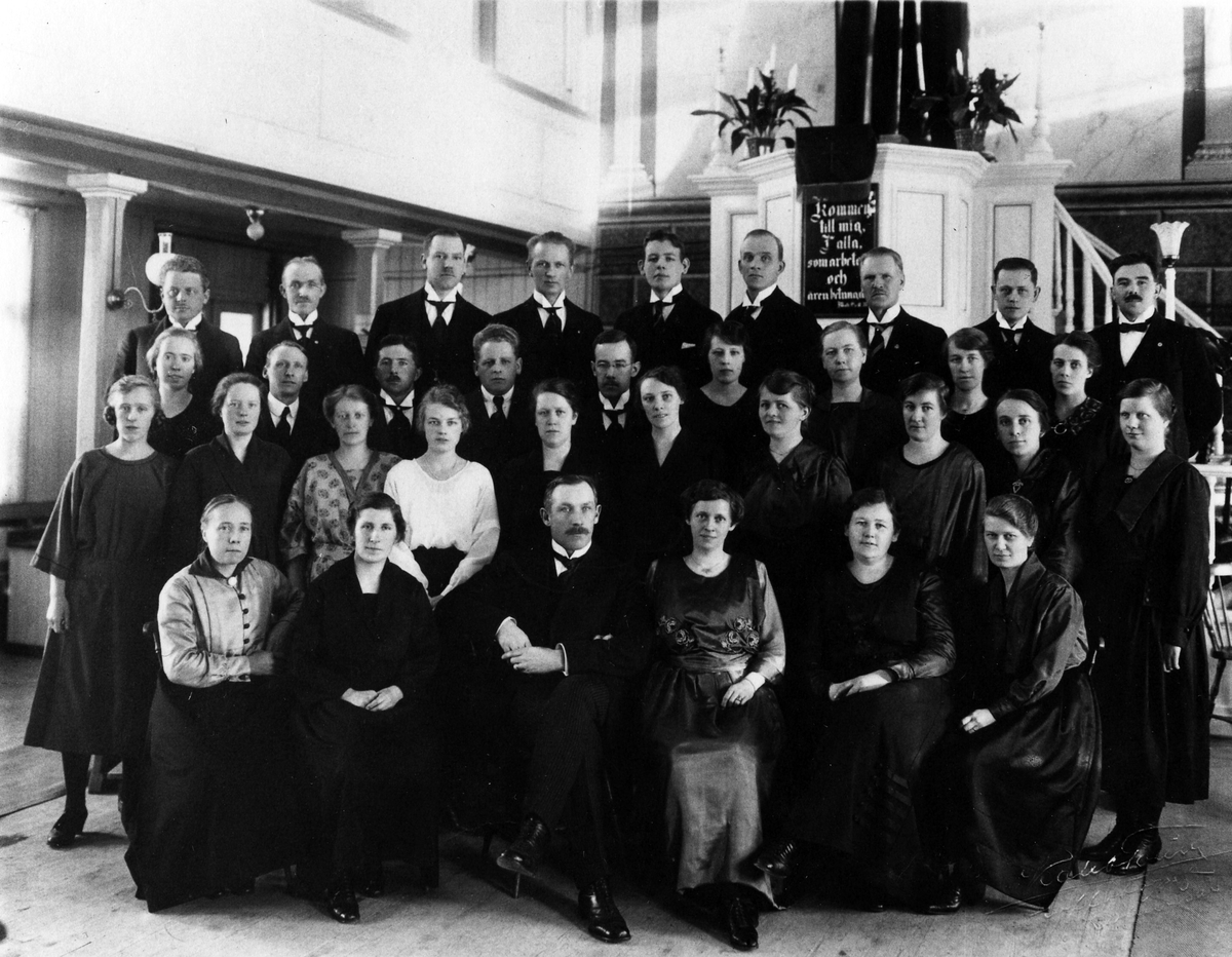 Gruppbild på 34 kvinnor och män i missionskyrkans kyrksal, stående och sittande, framför kyrkans predikstol. Under antependiet en tavla med ett bibelcitat.
