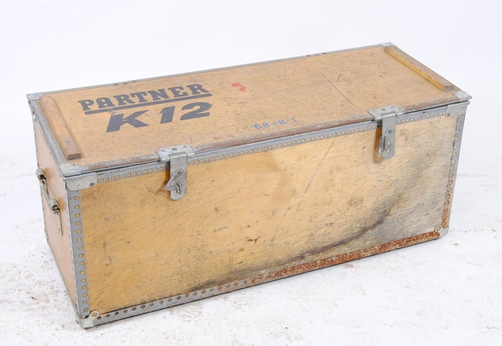 Transportlåda av plywood med plåtskodda hörn. På locket står det "PARTNER K12" tryckt i svart samt med blå schablonmålad text "6B-8-1".