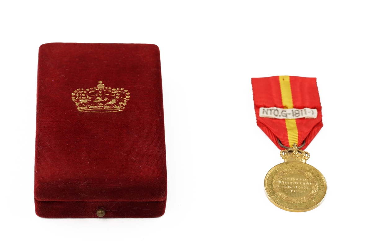 Gullmedalje med krone festet i et tekstilbånd med festenål liggende i etui