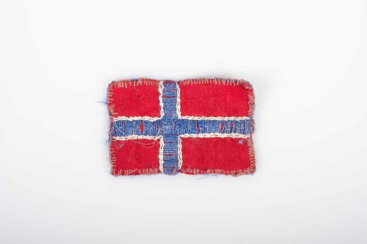 Et lite norsk flagg. Den er sydd og brodert på hånd. Den er sydd sammen av to stoffbiter, rød på forsiden og blå på baksiden. Det blå og hvite krysset på flagget er brodert.