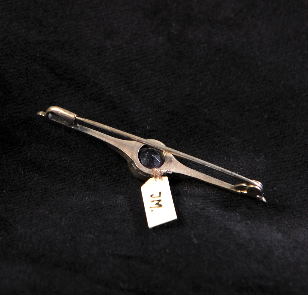 En brosch av silver (?) i form av en nål med infattad lila glassten (ska efterlikna ametist), med markasiter vid sidorna. Slitna stämplar.