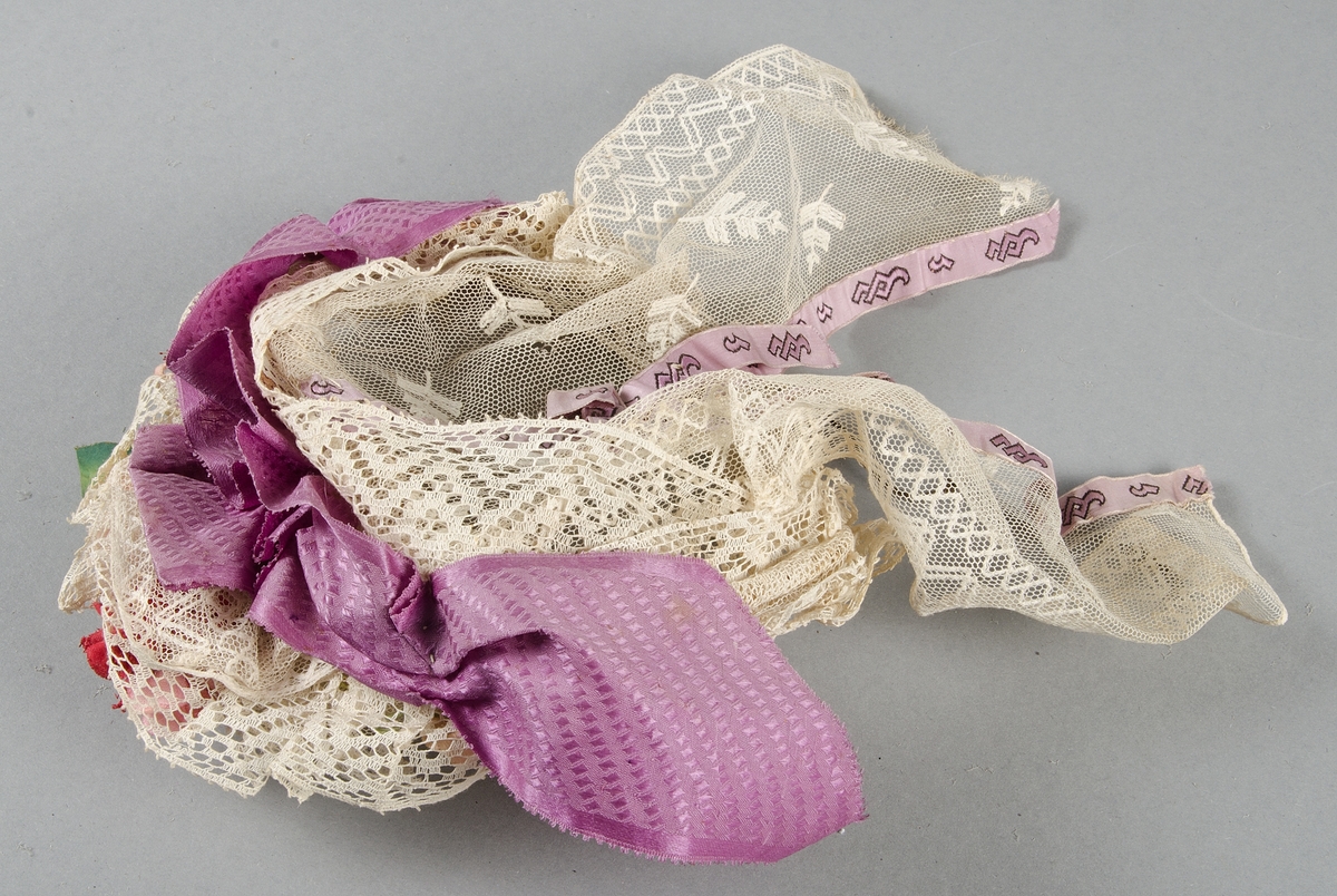 Hårklädsel av vita spetsar, mönstervävda lila band och rosa och röda tygblommor på stomme av mässing-tråd (?). Spetsarna, banden och tygblommorna smutsiga.