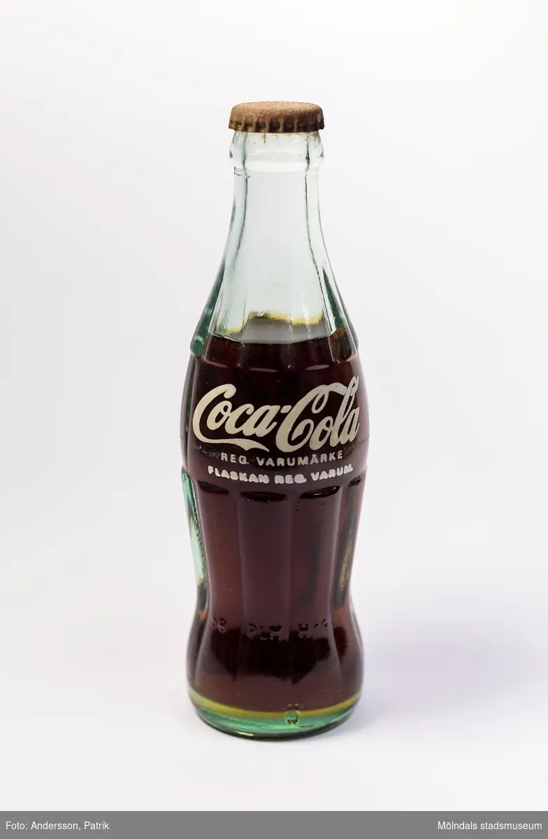 Coca-Cola i glasflaska från slutet av 1980-talet. Flaskan är tillverkad i grönaktigt glas.
Den 23 april 1985 ändrade Coca-Cola receptet på sin dryck som därmed fick namnet "New Coke". Det nya receptet blev ett totalt fiasko. Efter påtryckningar från konsumenterna återupptogs därför det gamla receptet redan i juli samma år, men då under namnet "Coca-Cola Classic". När "Coca-Cola Classic" kom tillbaka, sålde den redan från början mer än "New Coke". 

Glasflaskans form är inspirerad av kakaobönan. Det geniala med flaskan är dess greppvänliga form med dess särpräglade utseende som gör att man känner igen den även om den greppas i totalt mörker. Glaset splittrades heller inte om flaskan tappades i marken. I början tillverkades flaskorna i färgerna blått, grönt och glasklart glas.

Glasingenjören Alexander Samuelson som formgav flaskan har koppling till Sverige. Han föddes nämligen i Kungälv den 4 januari 1862. Under sina ungdomsår arbetade han på Surte glasbruk. Vid 21 års ålder flyttade han med sin storebror till USA, där han fortsatte att arbeta på olika glasbruk och blev så småingom chef. I början av 1910-talet anordnades en tävling för att designa en flaska till Coca-Colan. Tävlingen vanns av Alexander och hans glasbruk. 1915 tog Alexander Samuelson patent på denna geniala utformning av Coca-Colaflaskan, en av världens mest välkända varuförpackningar.

Eftersom livsmedelslagen i Sverige hade förbud mot koffein och fosforsyra, kunde inte Coca-Colan börja säljas i Sverige förrän 1953 då förbudet upphäves. Det var då Pripps bryggerier som tog hem lincensen för att börja tillverkningen av den amerikanska drycken i Göteborg, Malmö och Sundbyberg. Coca-Colan hade först svårt att konkurrera med storheter som Sockerdricka, Champis, Festis och Zingo. Det var först i slutet av 1970-talet som försäljningen ökade ordentligt.

Enligt inkommen kommentar: Enligt inskriften är flaskan tillverkad 1968 på Hammars glasbruk. Men drycken kan ju vara från 1980-talet.