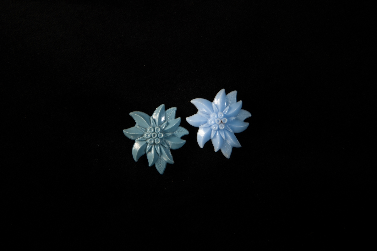 Ett par (?) clipsörhängen i plast, i form av en blå blomma. Örhängena har olika nyans av blå.