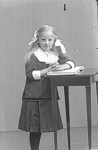 Aina Larsson, en liten flicka i kjol, med rosetter i håret stående lutad mot en pulpet med en uppslagen bok.