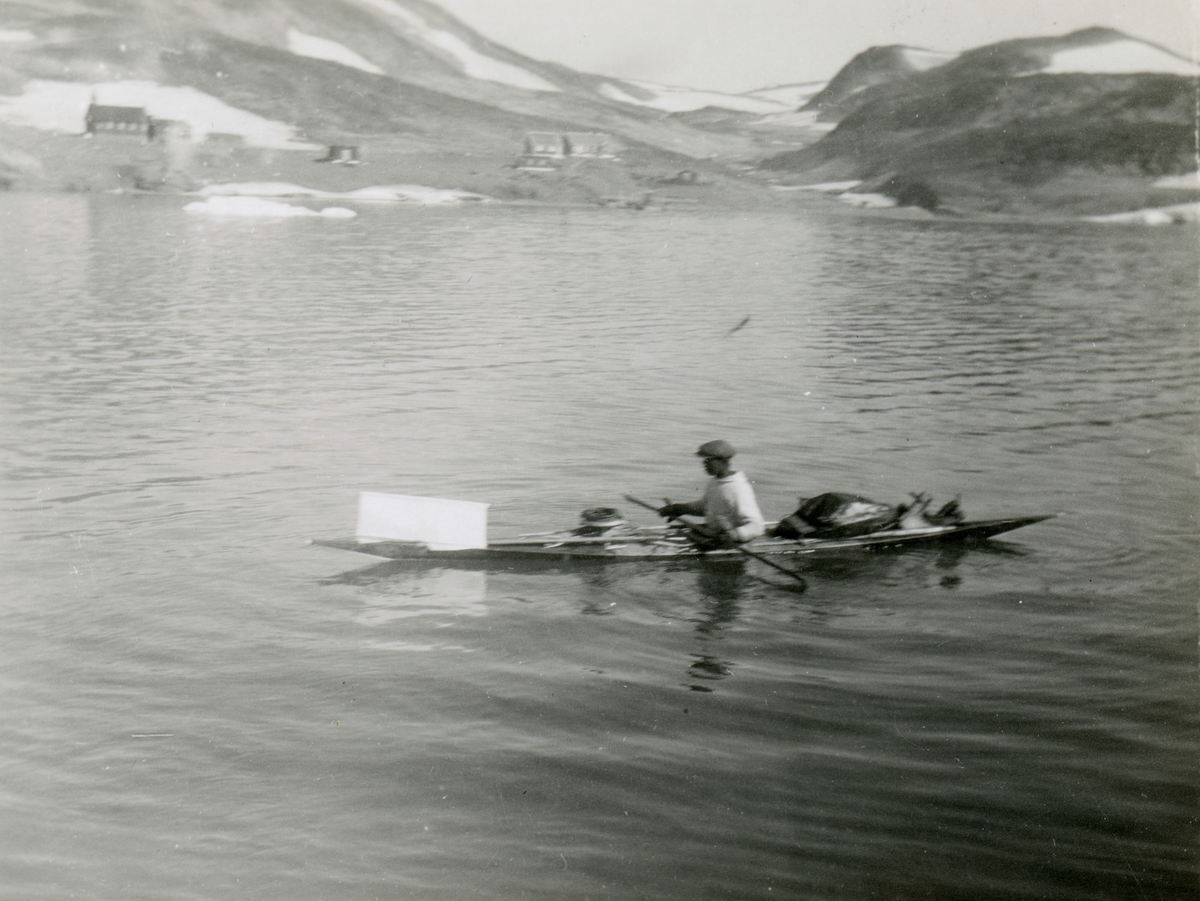 En mann fra Grønland er ute å padler i en kajakk med diverse utstyr ombord. Bebyggelse på land i bakgrunnen