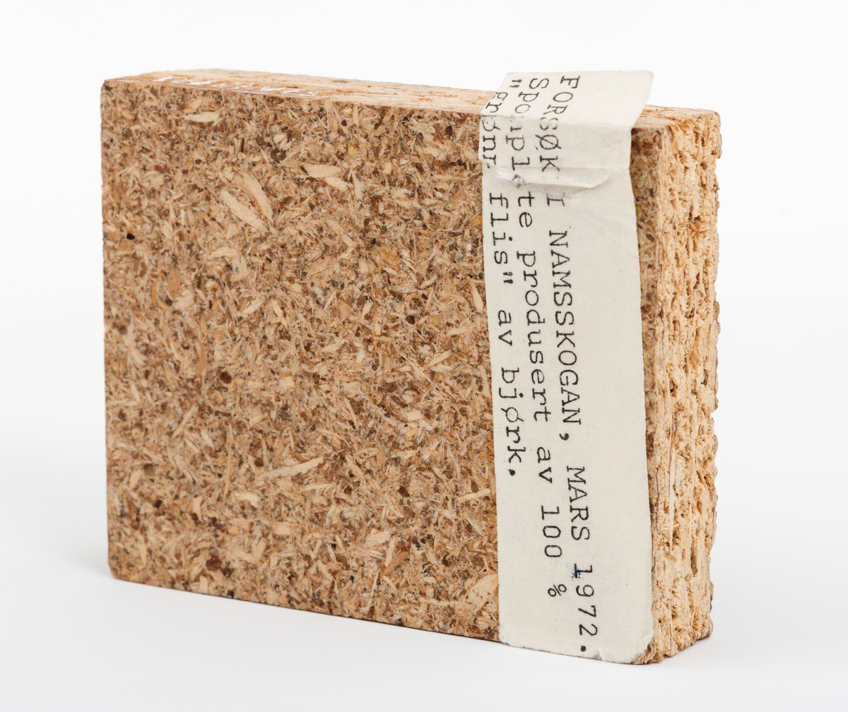 En rektangulær sponplate, vareprøve på 8,8 cm x 7 cm x 1,8 cm. Plata har glatt fram- og bakside.  På vareprøvas ene side er det påklistret en lapp med denne teksten: "Forsøk i Namskogan, mars 1972. Sponplate produsert av 100 % "grønn flis" av bjørk"