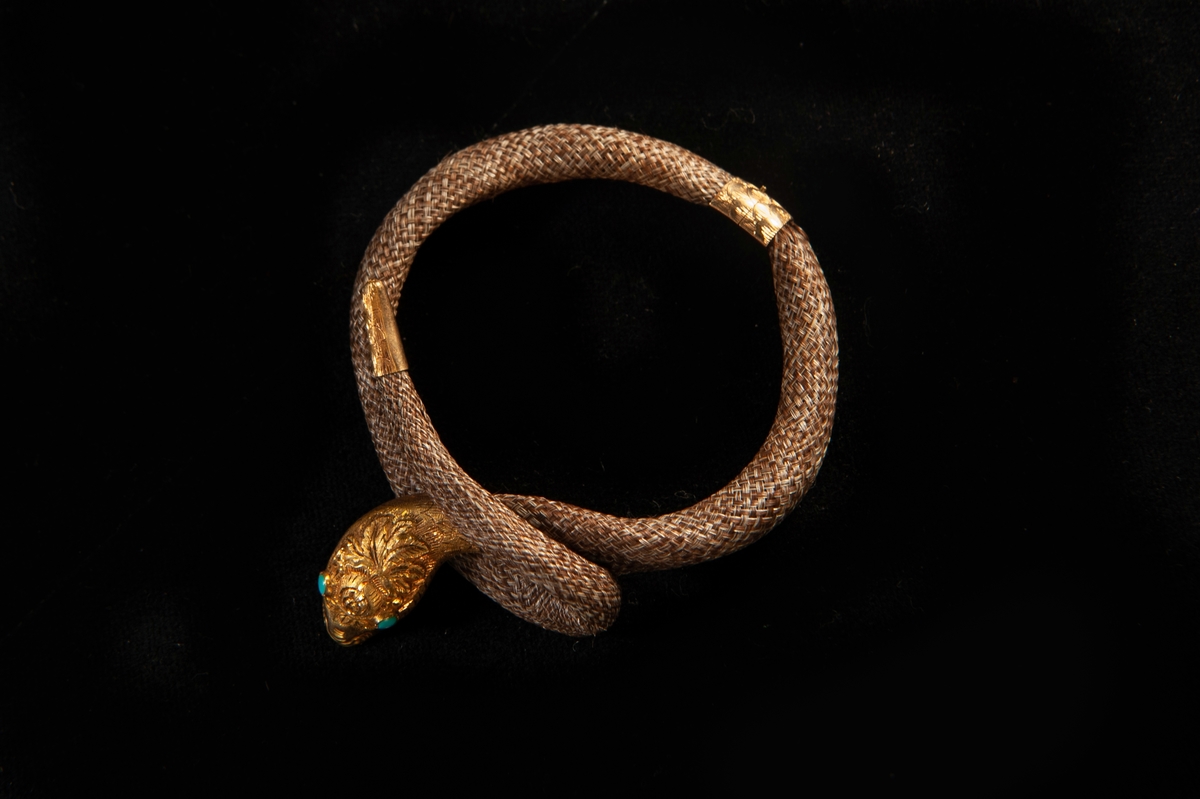Ett armband av cendré hår i flätat hårarbete. Formad som en orm med huvud, stjärt och lås i guld. Turkoser infattade som ögon. Graverad dekor på huvudet. Stämplar bak på huvudet.