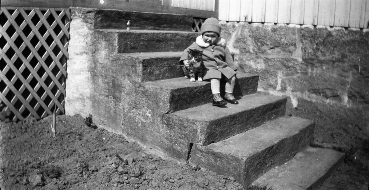 Ett litet barn sitter på en stentrappa med armen om en katt i solskenet. Barnet är klätt i mössa, pälskragad rock, strumpor och nätta kängor.