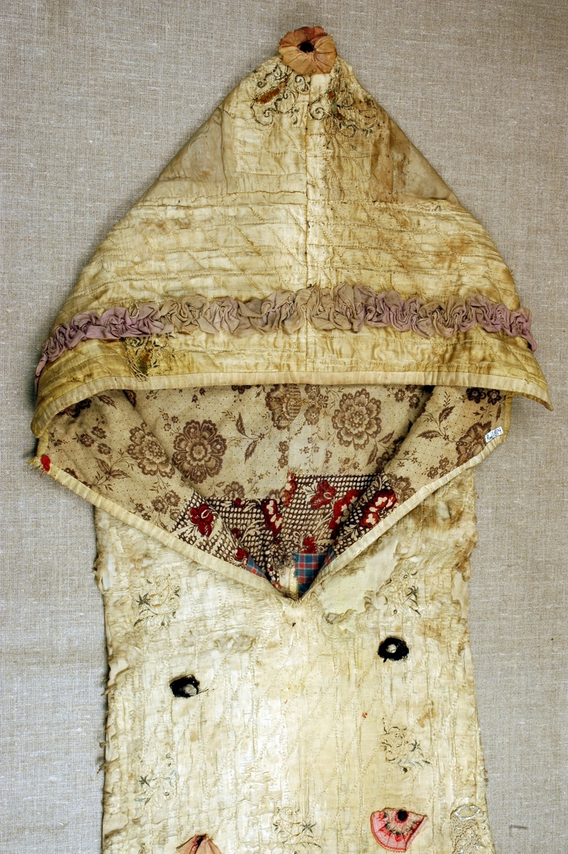 Dåpspose i brodert og vattert silke med stikninger, dekorert med blonde- og stoffrosetter med pynteknapper, perleblonde, rysjer og band. Foret med tre forskjellige stoffer, med ruter og blomster.