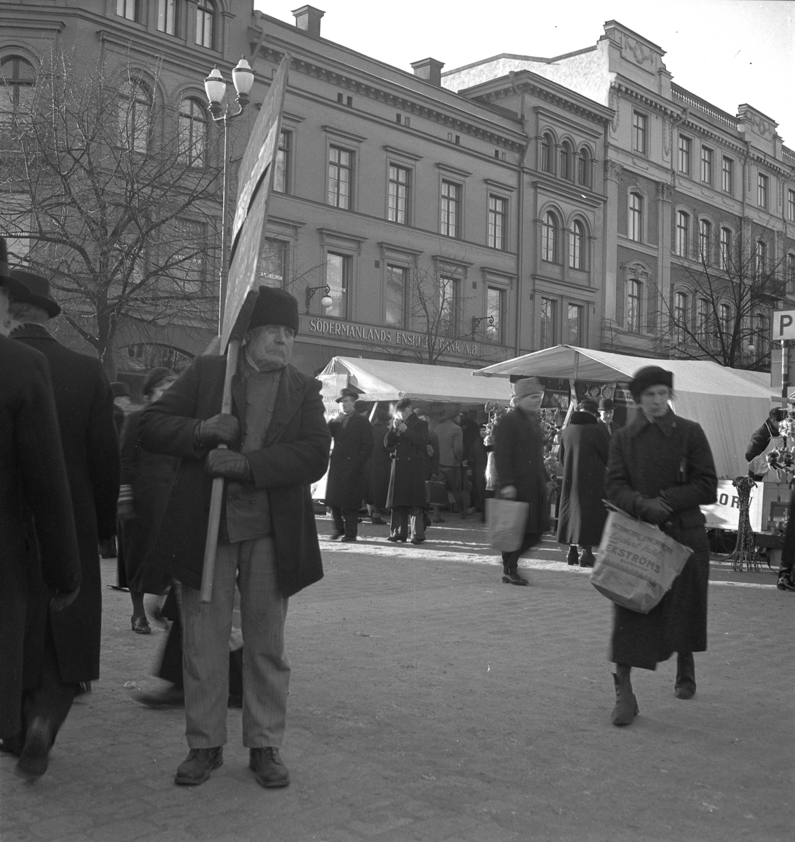 Hindersmässan i Örebro den 29 januari 1937. Folk vandrar mellan marknadsstånden på Stortorget. En man med ett plakat framträder i förgrunden.