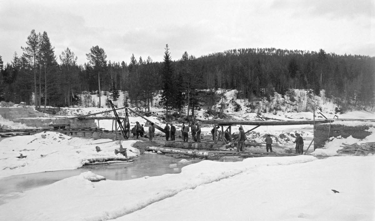 Anleggsarbeid ved Kjellåsfossen i Flisa, muligens i 1924.  Fotografiet er tatt vinterstid, da vannføringa var lav, slik at det var mulig å arbeide i elveløpet uten at vann og strøm skapte for store problemer.  Da dette fotografiet ble tatt var damarmene på begge sider av elveløpet ferdigmurte med kvadret naturstein som råstoff.  Anleggsarbeiderne holdt på midt i elveløpet der det skulle oppføres et damkar med et skråplan på motstrøms side (jfr. SJF-F.002262), slik at det ikke skulle bygge seg opp digre hauger av fløptingstømmer ved denne konstruksjonen.

Tidlig på 1900-tallet framsto Kjellåsdammen som et stort og kostbart fløtingsanlegg, som ble betjent av to damvoktere gjennom hele fløtingssesongen.  Hovedpoenget med anlegget var å magasinere vann, som kunne porsjoneres ut, slik at det ble mulig å få tømmeret videre nedover i retning Glomma også når vannstanden sank.  Effekten av dammen var omdiskutert, og enkelte mente at anlegget kunne fjernes, så snart det var utført litt mer mineringsarbeid i det nedenforliggende elveløpet.  I stedet bestemte fløtingsledelsen å bygge en ny og solidere Kjellåsdam, en murt steindam.  Arbeidet med denne konstruksjonen pågikk i perioden 1916-1924.  Kjellåsdammen var en såkalt attholdsdam, som skulle kunne brukes både til å heve vannspeilet i den ovenforliggende delen av elveløpet og til å regulere vannføringa i den nedenforliggende delen av Flisa-elva.
