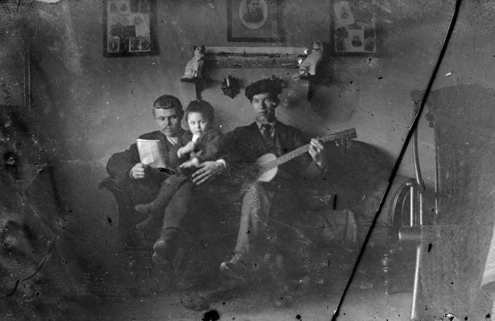 Leirfjord. En lesende mann med barn på fanget, og en mann med gitar. Alle fotografert i en sofa inne i en stue.