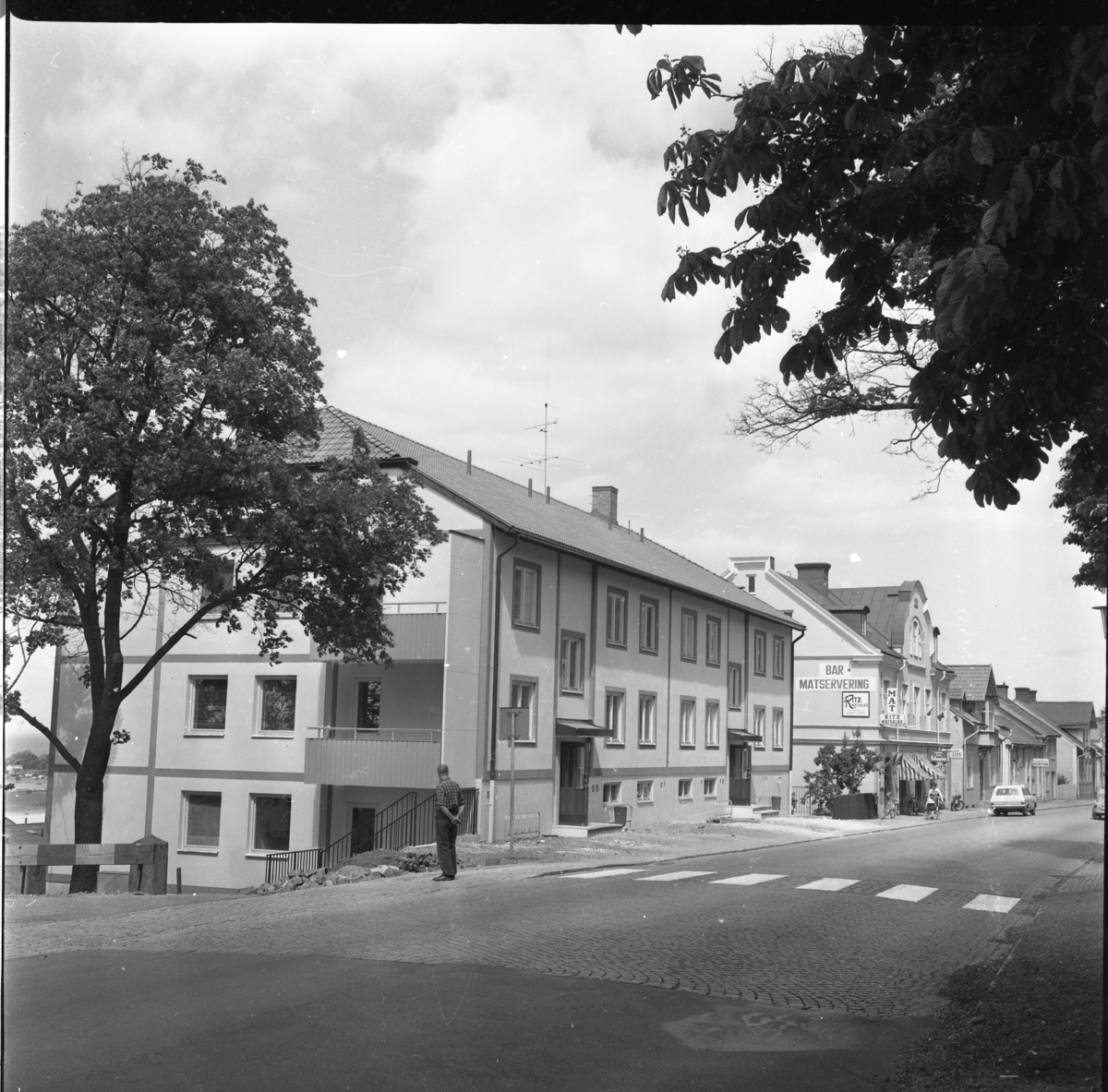 Nybyggt hyreshus på Brahegatan 1 i Gränna. Längre ner på gatan skyltas för Ritz Matsalar, mat och polkagrisar. En man i rutig skjorta och tofflor tittar mot huset.