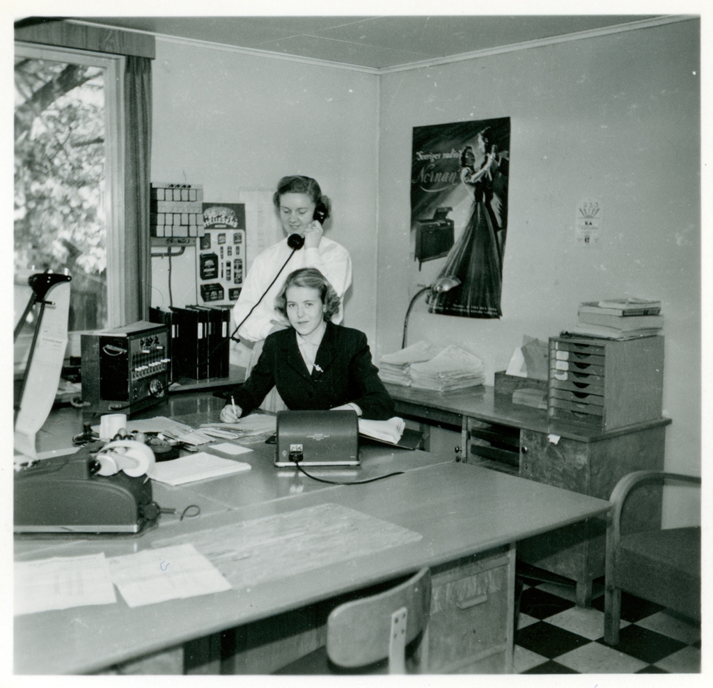 Två av kontorspersonalen på Monarks kontor. Den ena kvinnan står och talar i telefon, Gunnel Andersson sitter ner och tittar in i kameran. Bakom dem på väggen sitter en affisch med reklam för Monarks radiomöbel "Nornan".