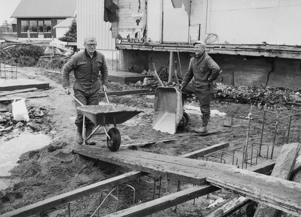 Leirfjord. Byggevirksomhet. To menn med trillebårer på byggeplassen. Bjarne Danielsen med trillebår og Henry Tømmervik står og ser på.
Finnes som VBH.F.2011-030-0032